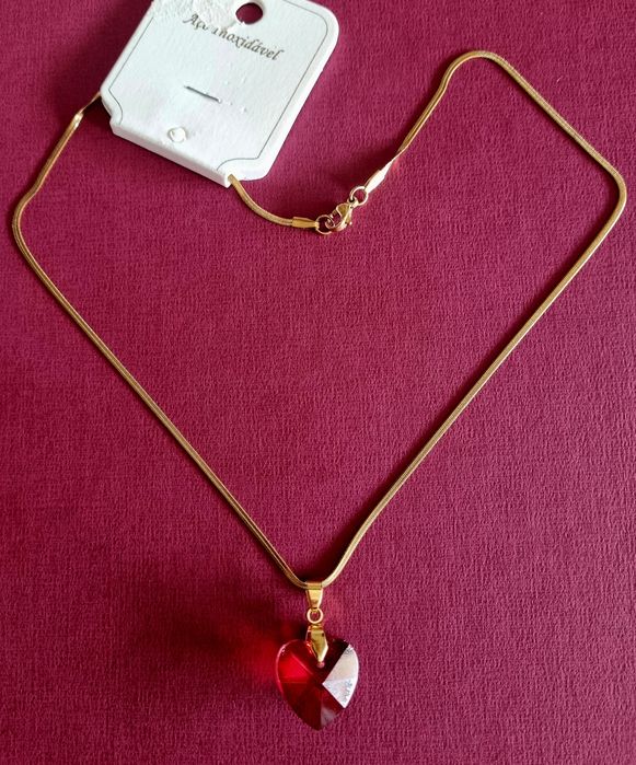 Colar dourado, em aço, malha fita e com um coração vermelho.