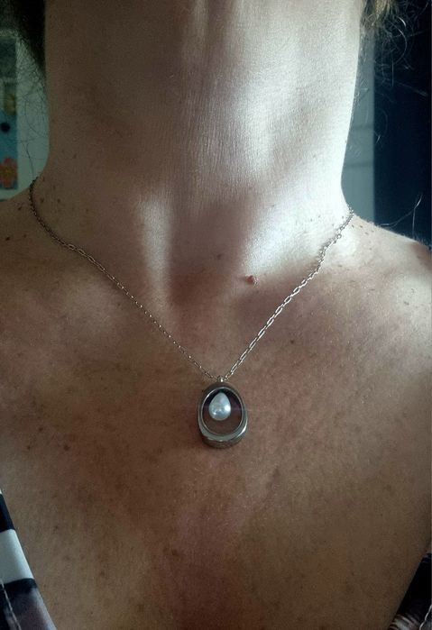 Colar prateado, em aço, com pingente oval e 1 pérola no centro. Tem extensor