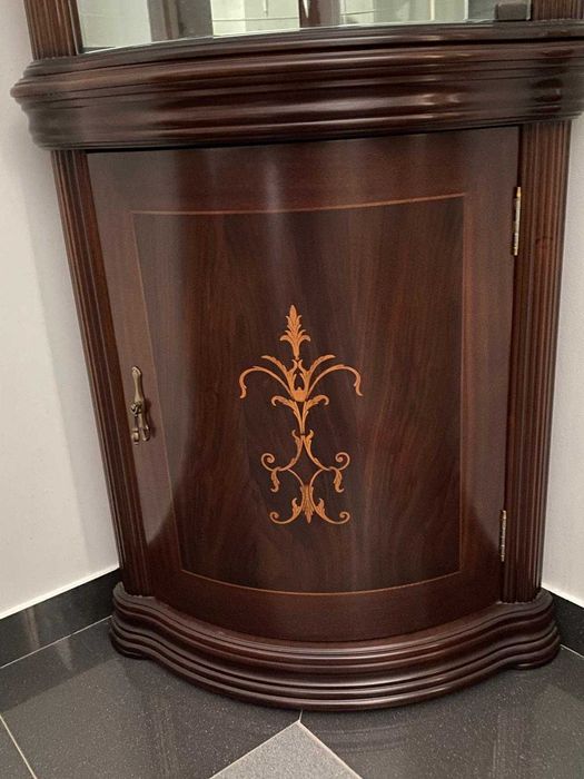 Vitrine de canto em madeira escura com painéis de vidro curvo e fundo espelhado.