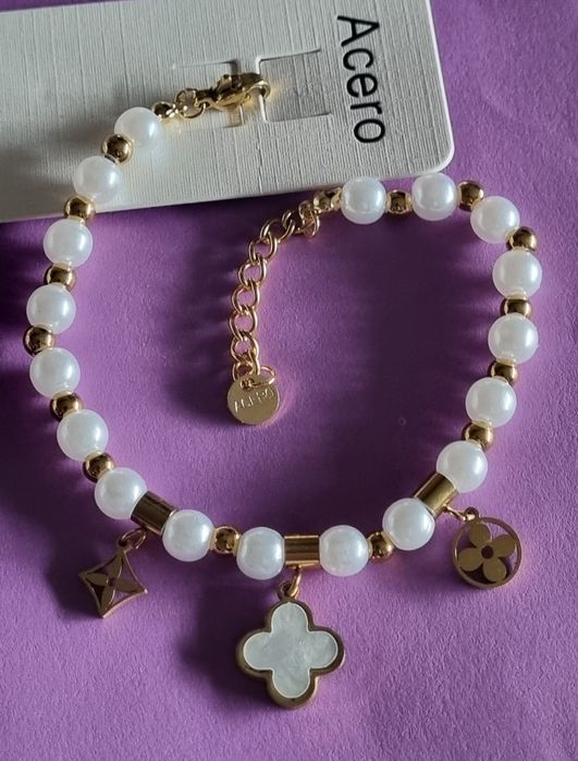 Pulseira dourada, em aço, com pérolas e um trevo branco. Tem extensor