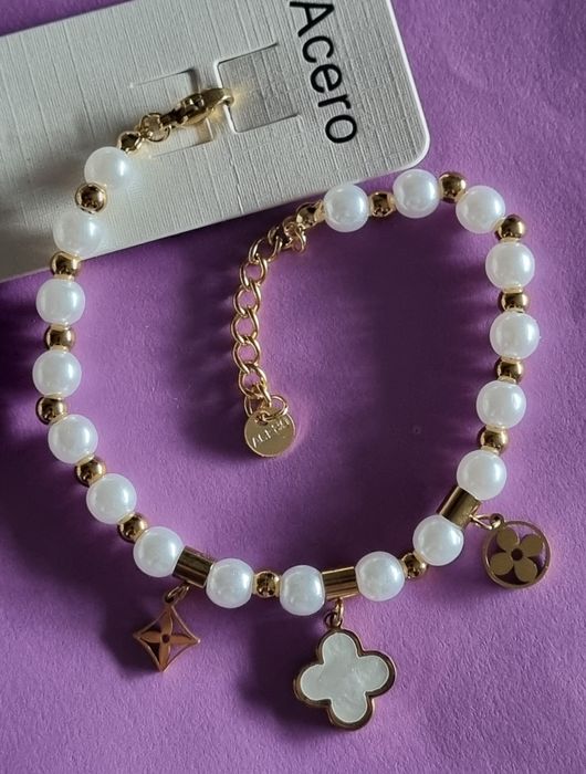 Pulseira dourada, em aço, com pérolas e um trevo branco. Tem extensor