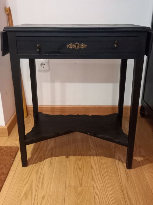 Mesa de apoio preta, antiga. Tem 2 abas pequenas e uma gaveta.