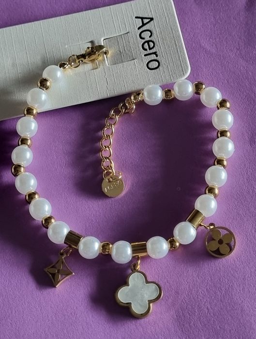 Pulseira dourada, em aço, com pérolas e um trevo branco. Tem extensor