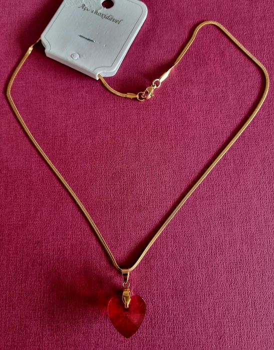 Colar dourado, em aço, malha fita e com um coração vermelho.