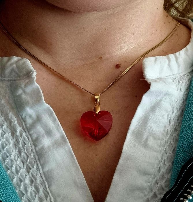 Colar dourado, em aço, malha fita e com um coração vermelho.