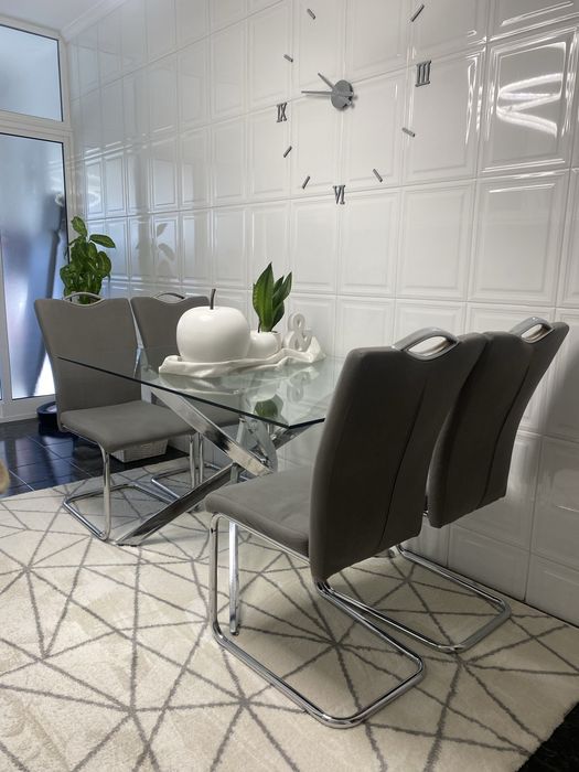 Esta é uma mesa de jantar moderna com um tampo em vidro transparente e uma elegante estrutura de pernas cruzadas em aço cromado brilhante.