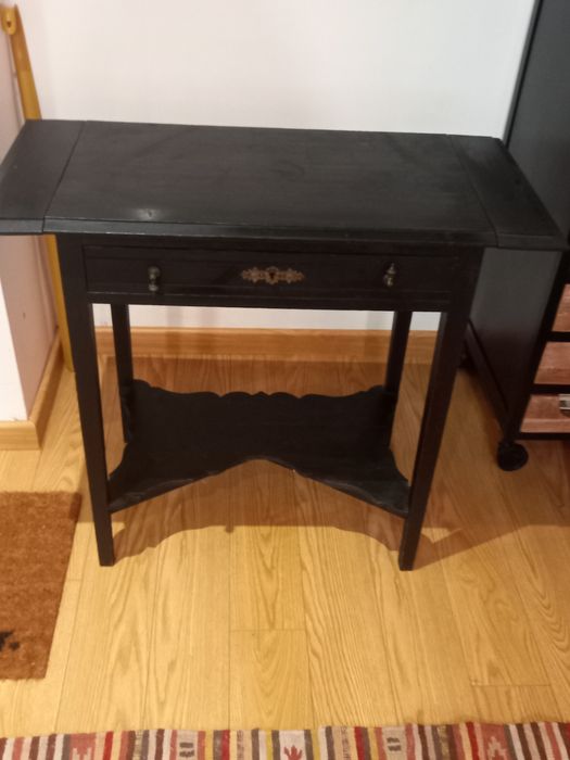 Mesa de apoio preta, antiga. Tem 2 abas pequenas e uma gaveta.