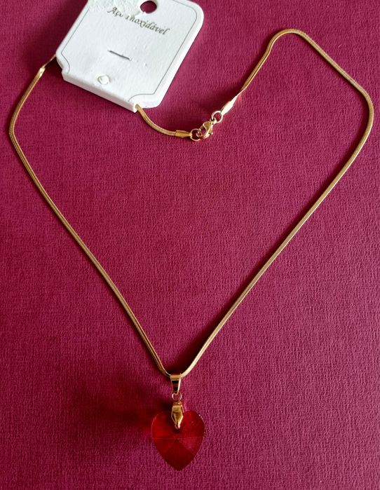 Colar dourado, em aço, malha fita e com um coração vermelho.