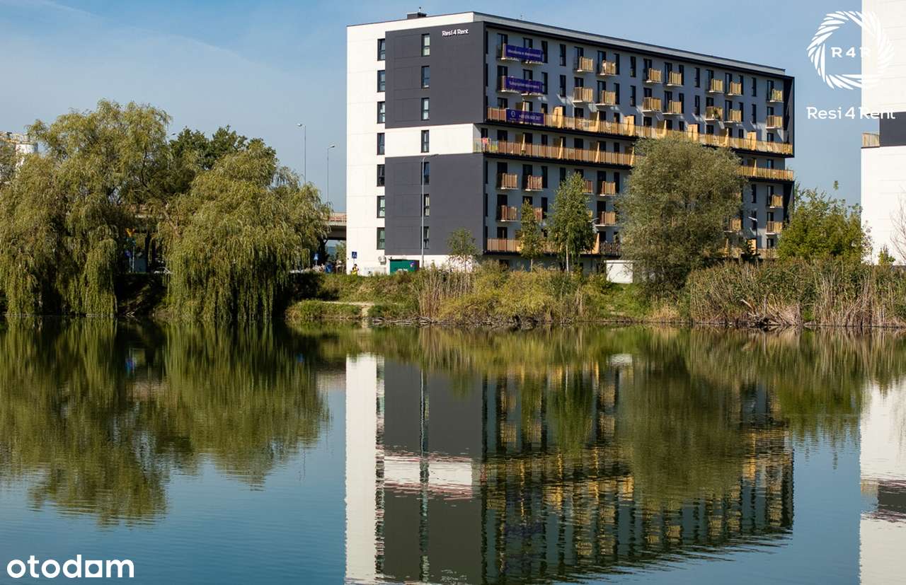 Nowoczesny budynek mieszkalny Resi4Rent nad wodą, z balkonami, dużymi oknami i zielonym otoczeniem, widoczny w spokojnej lokalizacji z odbiciem fasady w jeziorze.