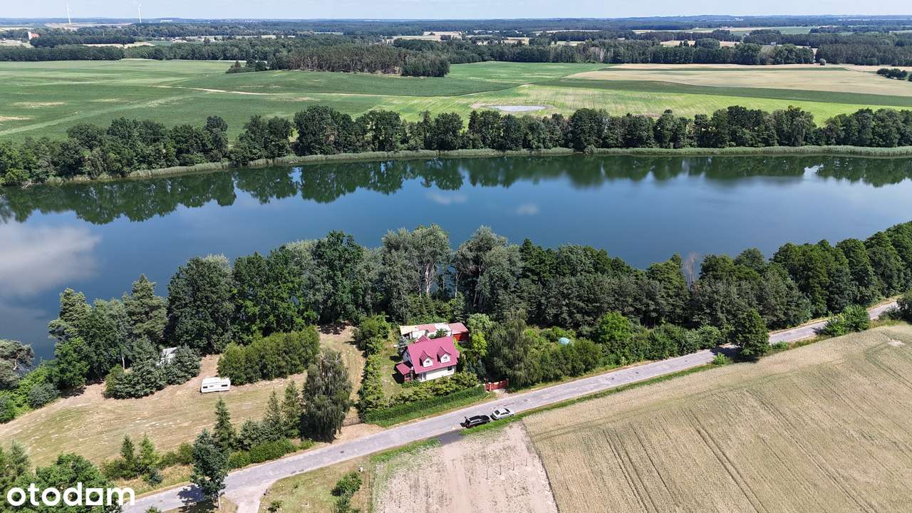 Dom z czerwonym dachem między drzewami nad Jeziorem Głęboczek Duży, pas wody wśród pól