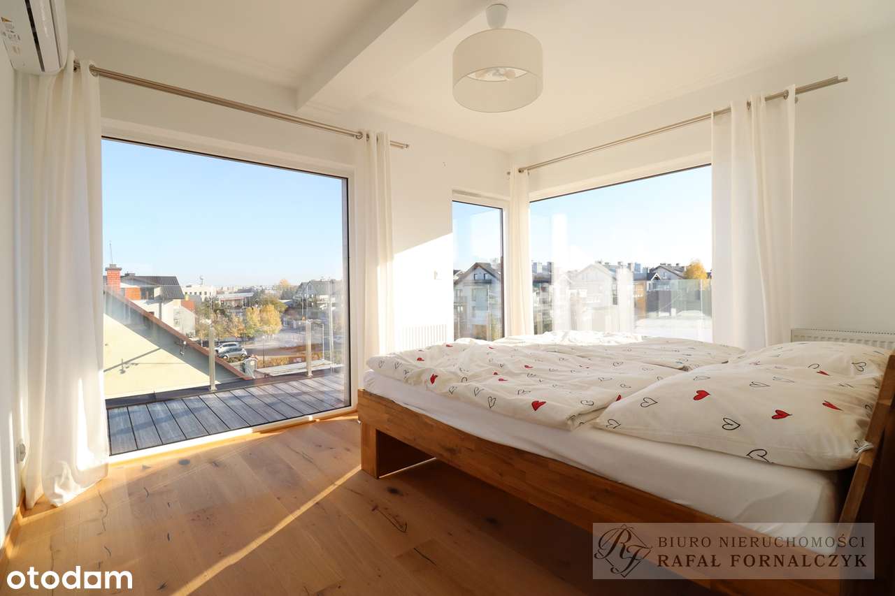 Przestronny taras apartamentu z drewnianą podłogą, szklanymi balustradami i leżakami, z widokiem na spokojną zabudowę miejską w słoneczny dzień.