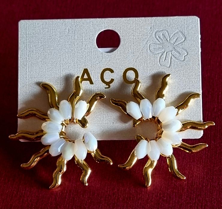 Brincos dourados, em aço, em forma de sol com pedrinhas brancas.  De pico.