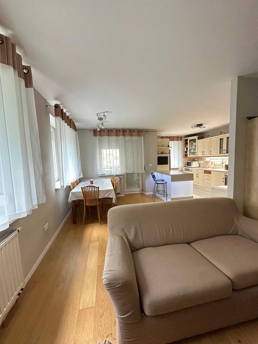 Jasny salon z aneksem kuchennym i częścią jadalnianą, wyposażony w beżową sofę, drewniany stół z krzesłami, duże okna z firanami oraz zabudowaną kuchnię w ciepłym, zadbanym standardzie.