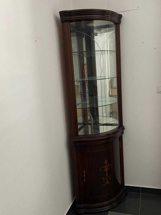 Vitrine de canto em madeira escura com painéis de vidro curvo e fundo espelhado.