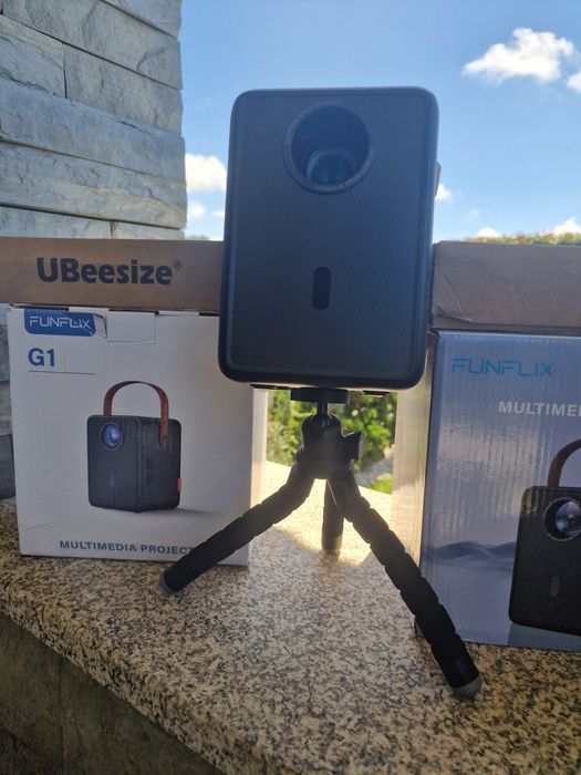 projetor portátil FunFlix G1 com alça de transporte.O dispositivo possui conectividade Wi-Fi 5G oferta de tripé