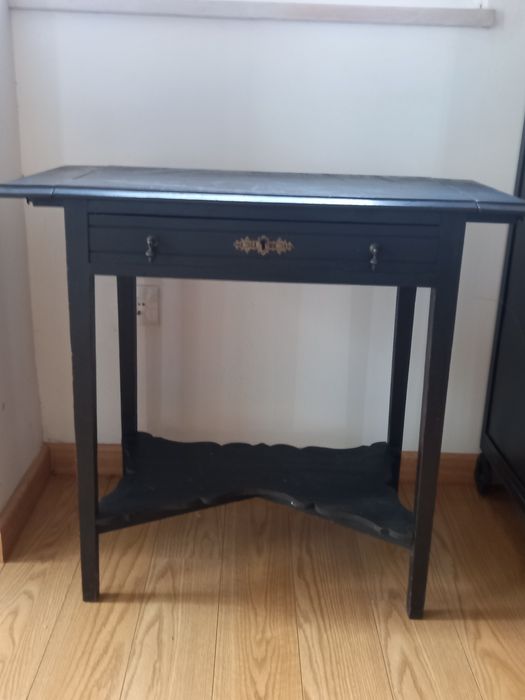 Mesa de apoio preta, antiga. Tem 2 abas pequenas e uma gaveta.
