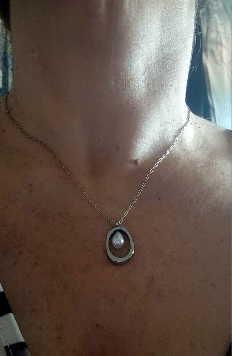 Colar prateado, em aço, com pingente oval e 1 pérola no centro. Tem extensor