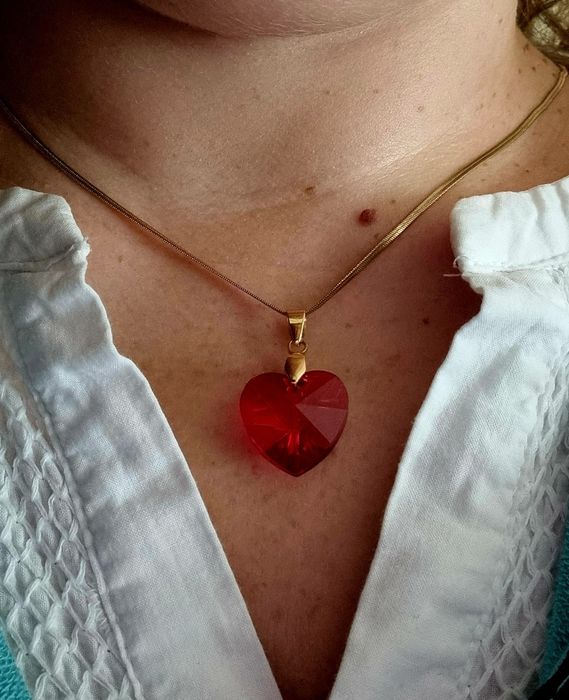 Colar dourado, em aço, malha fita e com um coração vermelho.