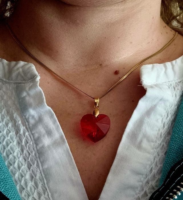 Colar dourado, em aço, malha fita e com um coração vermelho.