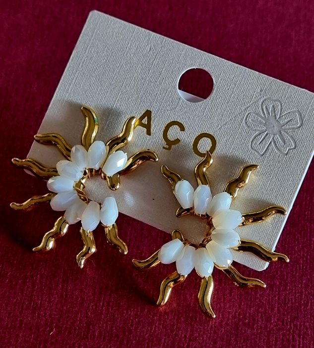 Brincos dourados, em aço, em forma de sol com pedrinhas brancas.  De pico.