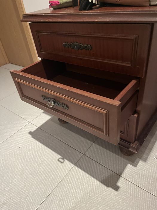 Mesa de cabeceira clássica de madeira escura com três gavetas e puxadores decorativos em metal escuro.