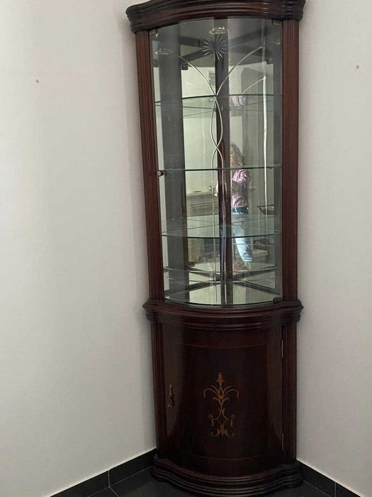 Vitrine de canto em madeira escura com painéis de vidro curvo e fundo espelhado.