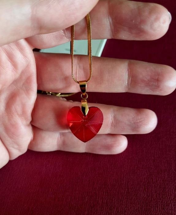 Colar dourado, em aço, malha fita e com um coração vermelho.