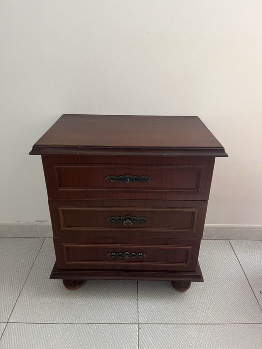 Mesa de cabeceira clássica de madeira escura com três gavetas e puxadores decorativos em metal escuro.