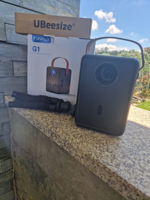 projetor portátil FunFlix G1 com alça de transporte.O dispositivo possui conectividade Wi-Fi 5G oferta de tripé