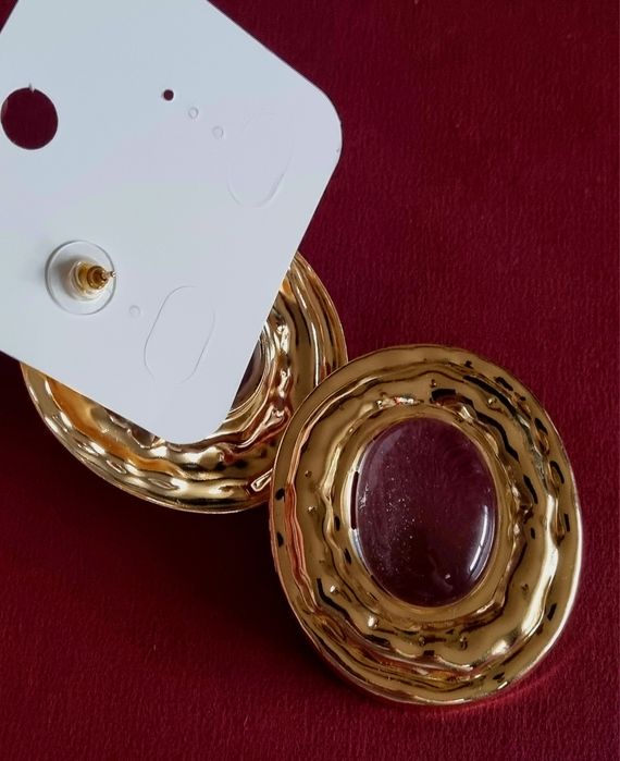 Brincos grandes dourados com acrílico oval transparente no centro.