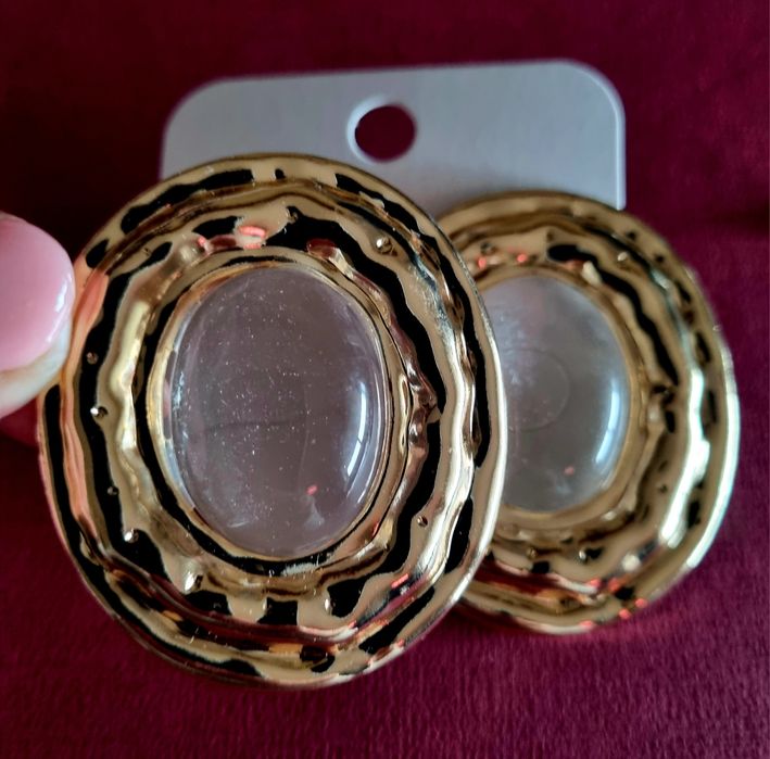 Brincos grandes dourados com acrílico oval transparente no centro.