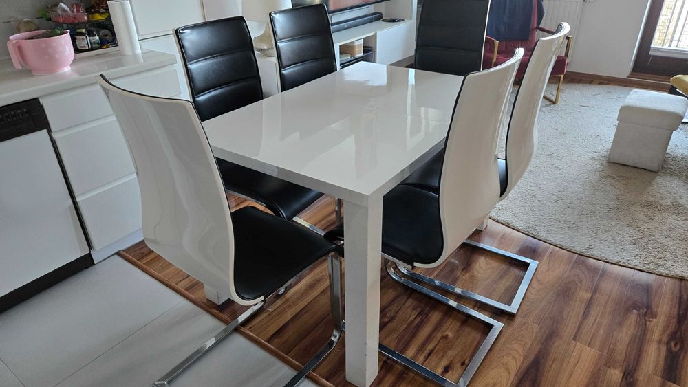 Biały rozkładany stół i 6 krzeseł z czarną tapicerką.