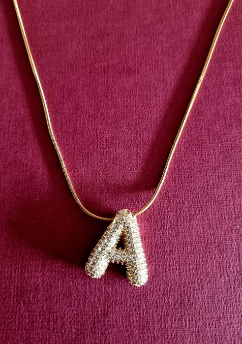 Colar dourado, em aço, com a letra "A" toda cravejada de zirconias.