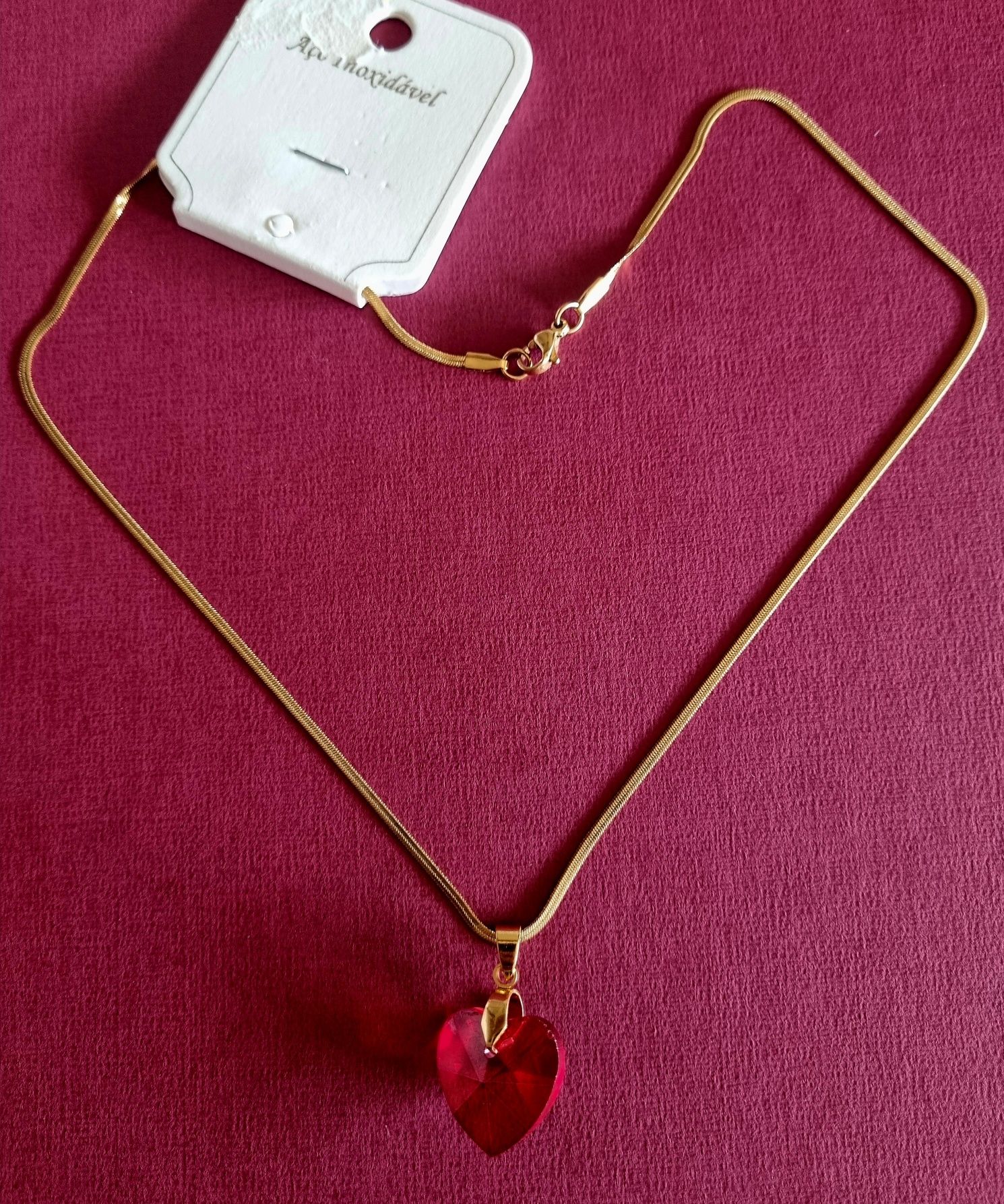 Colar dourado, em aço, malha fita e com um coração vermelho.