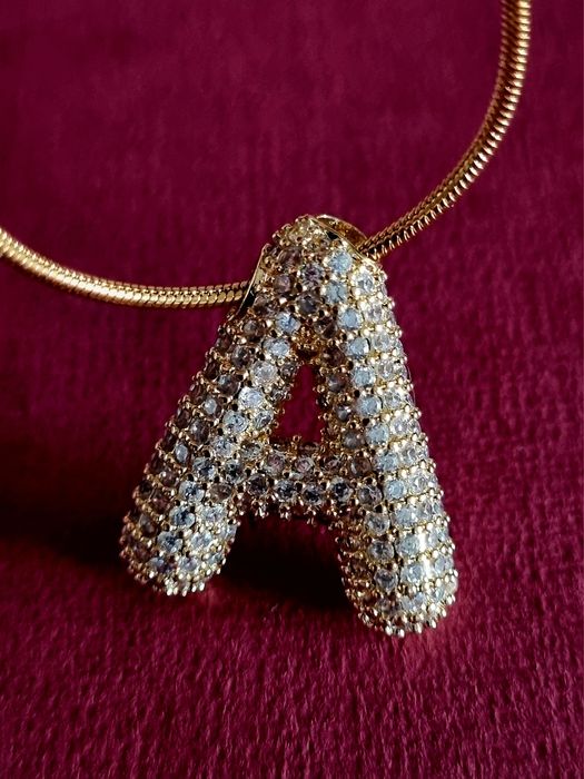 Colar dourado, em aço, com a letra "A" toda cravejada de zirconias.