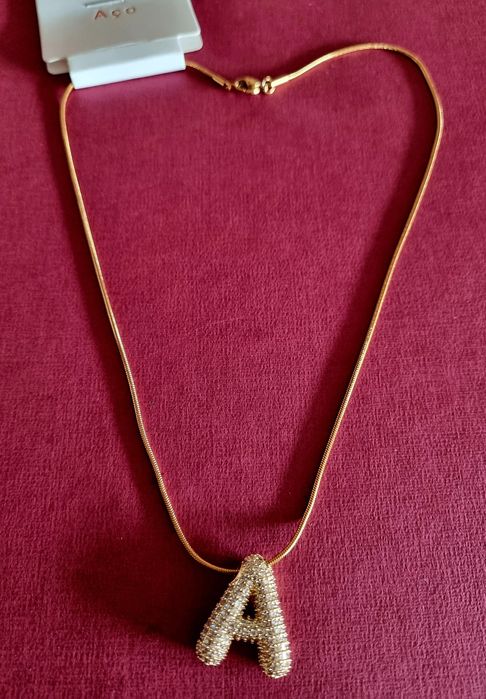 Colar dourado, em aço, com a letra "A" toda cravejada de zirconias.