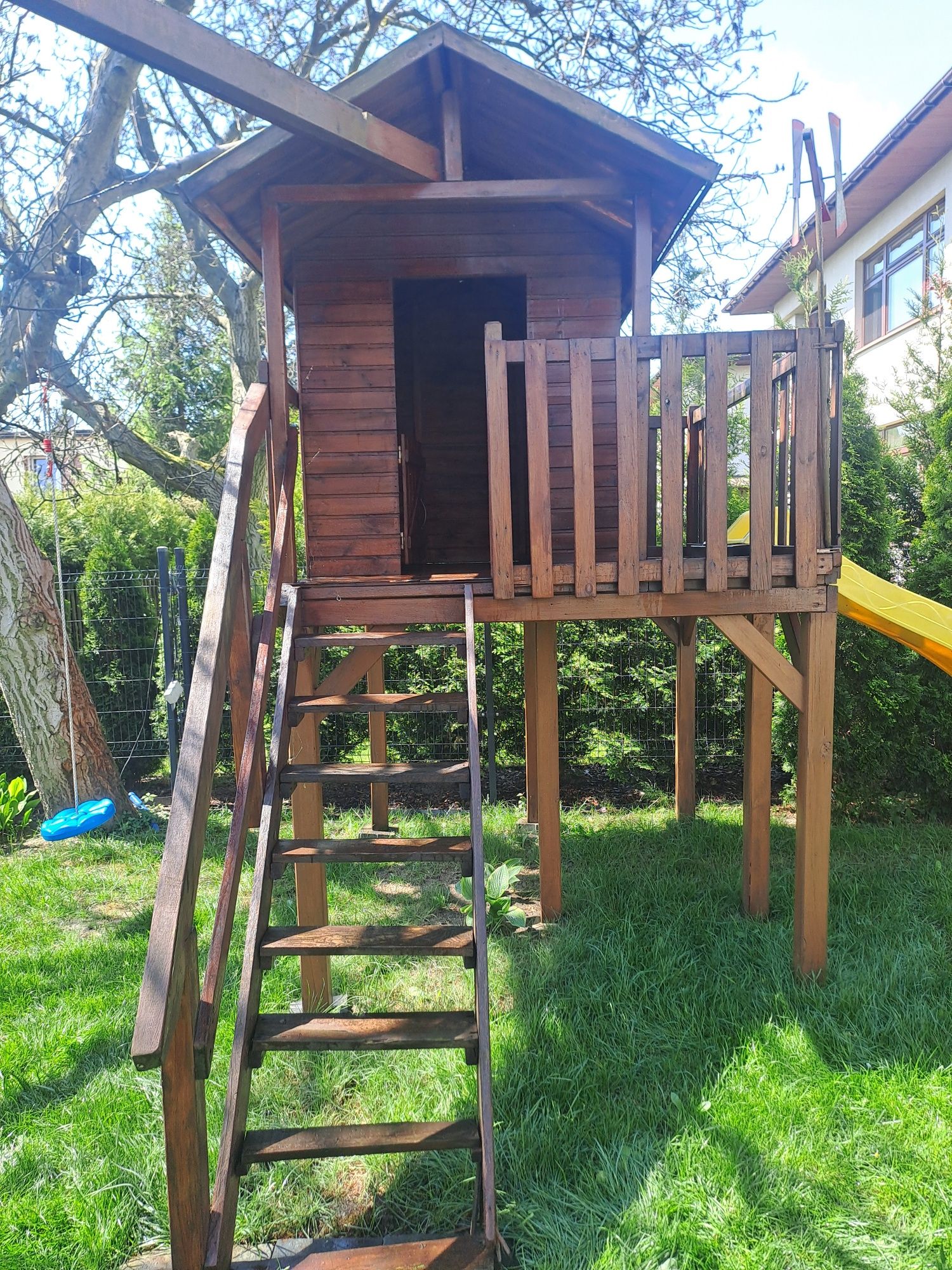 Drewniany domek dla dzieci na podwyższeniu ze schodami i zjeżdżalnią.