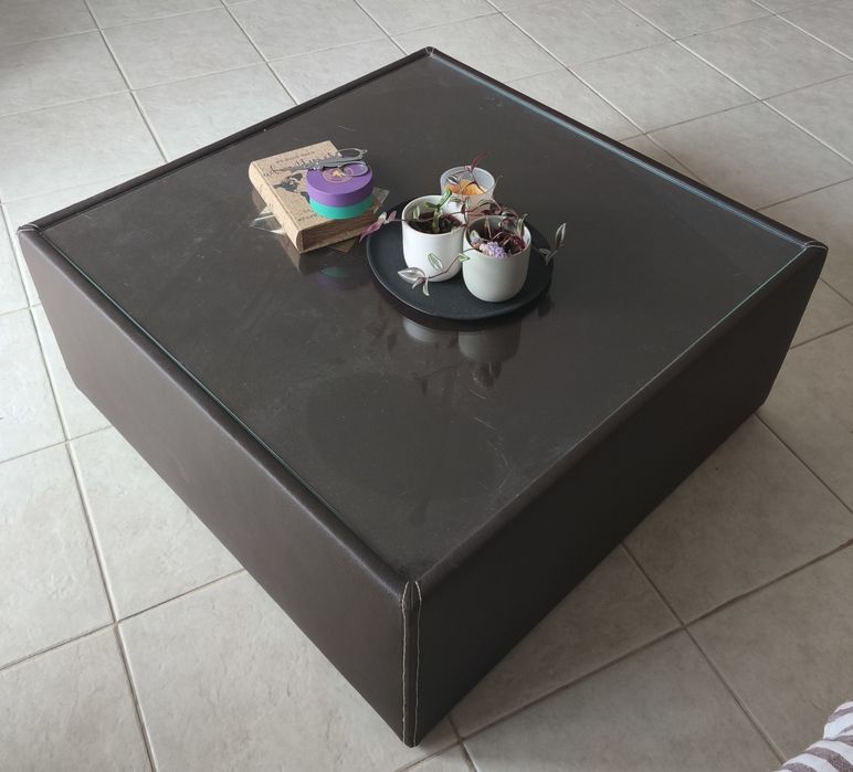 Mesa de centro baixa, quadrada, em pele sintética e tampo de vidro.