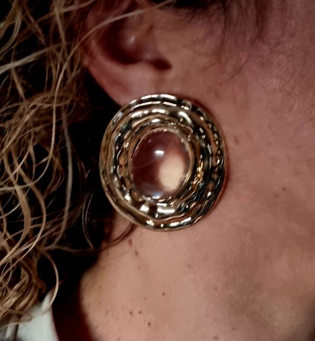 Brincos grandes dourados com acrílico oval transparente no centro.