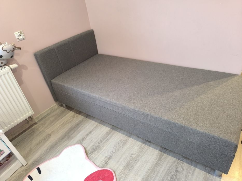 Tapczan, szare, tapicerowane łóżko jednoosobowe z wezgłowiem.