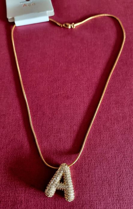 Colar dourado, em aço, com a letra "A" toda cravejada de zirconias.