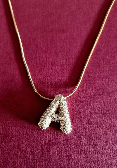 Colar dourado, em aço, com a letra "A" toda cravejada de zirconias.